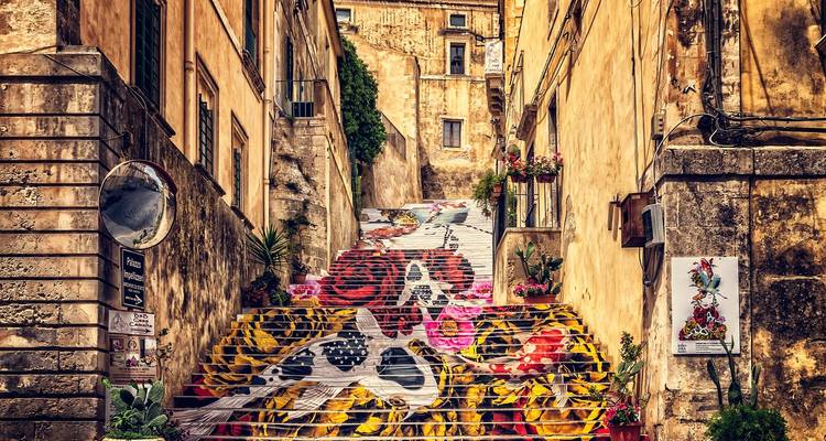 Narrow historic alleyway with wide stone staircase covered in colorful painted floral and fruit motifs between warm-toned buildings.