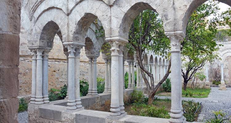 Peaceful cloister courtyard with stone arches, columns and a small garden of trees and greenery.