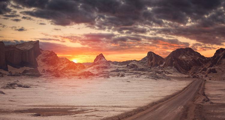 Uitgestrekt maanachtig woestijnlandschap bij zonsondergang met dramatische wolken en een eenzame zandweg die er kronkelend doorheen loopt.