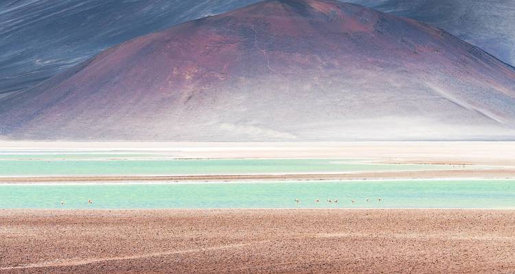 Turquoise en wit mineraal meer met roodbruine vulkanische kegel die erachter oprijst in hoog woestijnlicht.