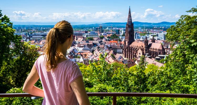 Junge Frau an einem Aussichtspunkt mit Blick auf Freiburgs Altstadt mit roten Dächern und Kathedrale an einem sonnigen Tag.