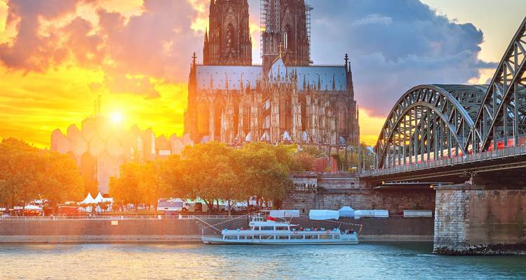 Coucher de soleil spectaculaire derrière la cathédrale de Cologne avec le pont Hohenzollern enjambant le Rhin et un bateau de croisière en contrebas.