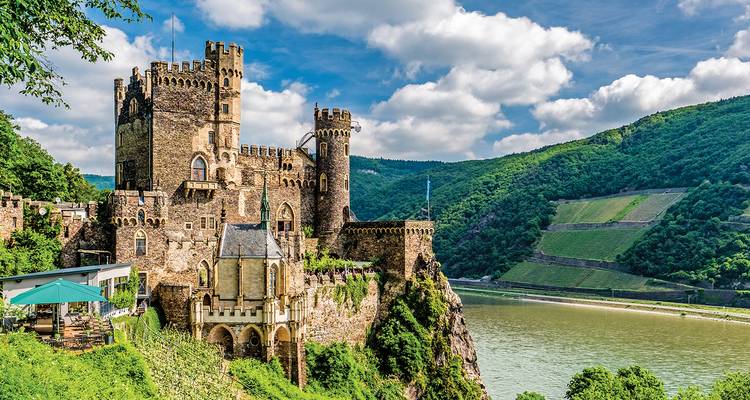 Château pittoresque de style Rheinstein perché sur une falaise au-dessus du Rhin avec des vignobles verts et un ciel bleu