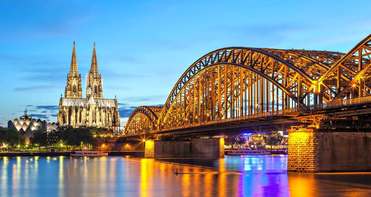Blue-hour panorama of the illuminated Hohenzollern Bridge and Cologne Cathedral reflecting on the Rhine.
