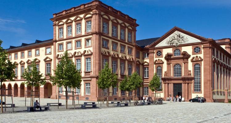 Le grand château baroque de Mannheim avec ses fenêtres symétriques fait face à une cour pavée où les visiteurs se détendent sur des bancs.