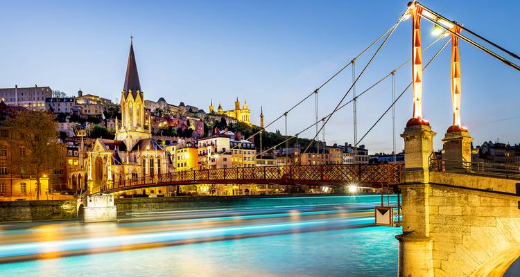 Escena nocturna de puente peatonal colgante iluminado e iglesia junto al río con reflejos borrosos del río.