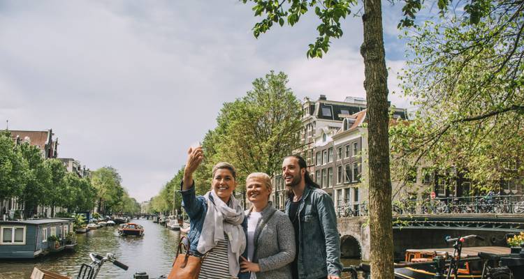 Trois amis prennent un selfie au bord d'un canal bordé d'arbres avec des maisons traditionnelles d'Amsterdam et des bateaux.