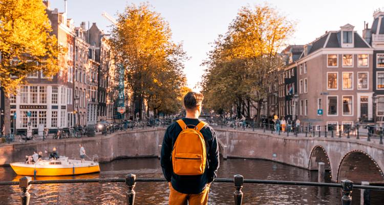 Un voyageur solitaire avec un sac à dos orange contemple un canal d'Amsterdam ensoleillé bordé d'arbres d'automne.