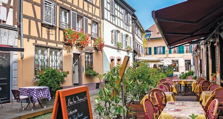 Charming narrow street lined with half-timbered Alsatian houses, colorful flowers and outdoor café tables under blue sky.