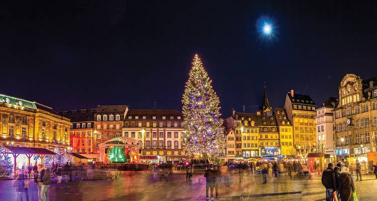 Scène de Noël nocturne à Strasbourg avec arbre illuminé et échoppes festives, flou de mouvement de la foule sous une lune brillante.