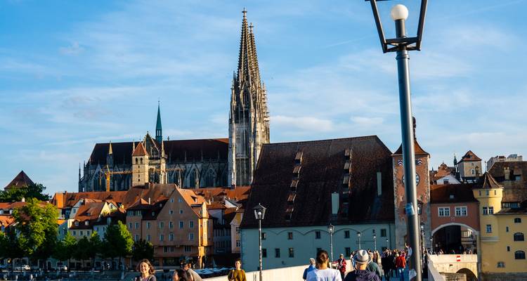 People stroll across Regensburg’s stone bridge toward the cathedral spires rising above colorful medieval rooftops.