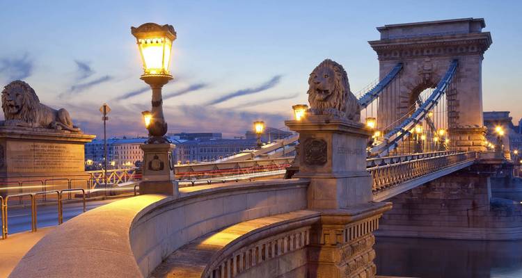 Vue nocturne du pont à chaînes de Budapest illuminé au-dessus du Danube avec des statues de lions et un ciel bleu tendre.