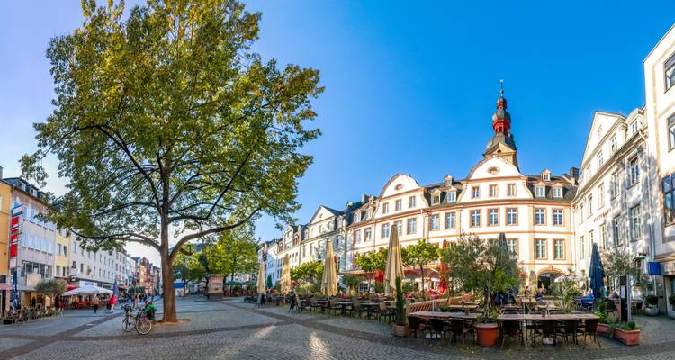 Place piétonne ensoleillée bordée de cafés et d'un bâtiment historique majestueux sous un ciel bleu éclatant.