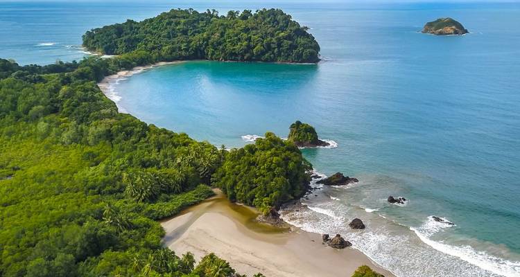 Vista aérea de una península verde y exuberante que abraza una bahía en forma de media luna de agua azul vívida con islotes distantes.