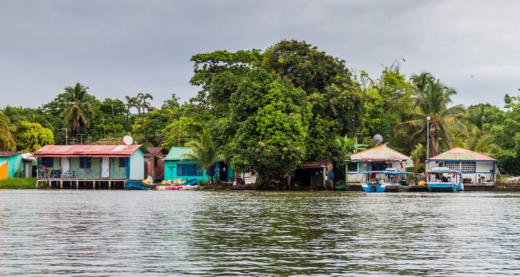 Coloridas casas de madera frente al río y pequeñas embarcaciones respaldadas por densa vegetación tropical en un día nublado.