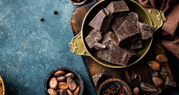 Bodegón rústico de trozos de chocolate negro, granos de cacao y platos de madera sobre una superficie de piedra azul.