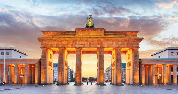 Porte de Brandebourg illuminée au crépuscule sous un ciel dramatique avec une place pavée vide devant.