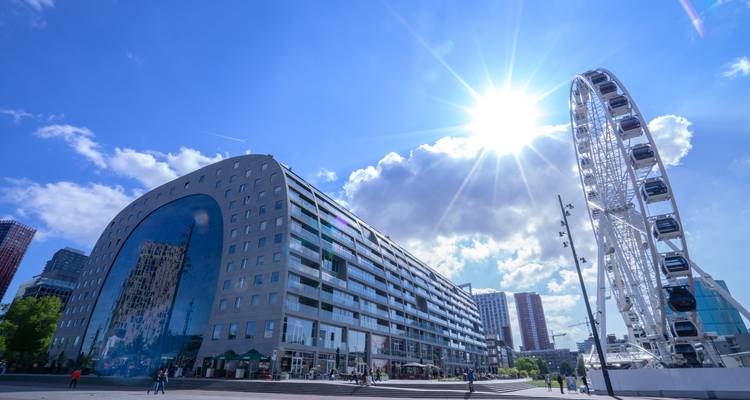 Moderne Markthal gebouw en reuzenrad in Rotterdam onder felle zonnestralen en blauwe lucht.