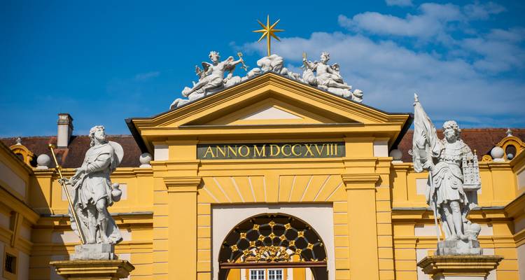 Nahaufnahme des verzierten gelben Eingangs der Stifts Melk mit Marmorstatuen und lateinischer Inschrift vor blauem Himmel.