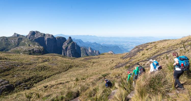 Des randonneurs descendent un sentier de montagne herbeux avec des aiguilles rocheuses spectaculaires et des vallées lointaines sous un ciel bleu dégagé
