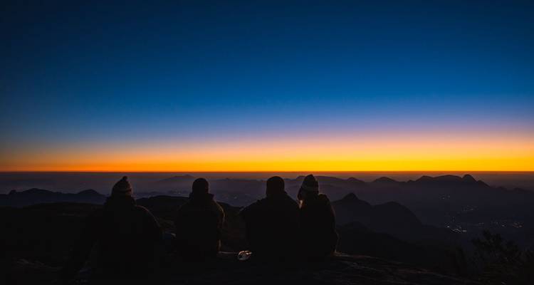 Quatre randonneurs sont assis sur une crête en regardant des couleurs orange et bleues éclatantes au-dessus des montagnes à l'aube