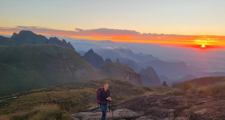 Randonneur avec bâtons de marche debout sur un affleurement rocheux surplombant des crêtes montagneuses spectaculaires au lever du soleil.