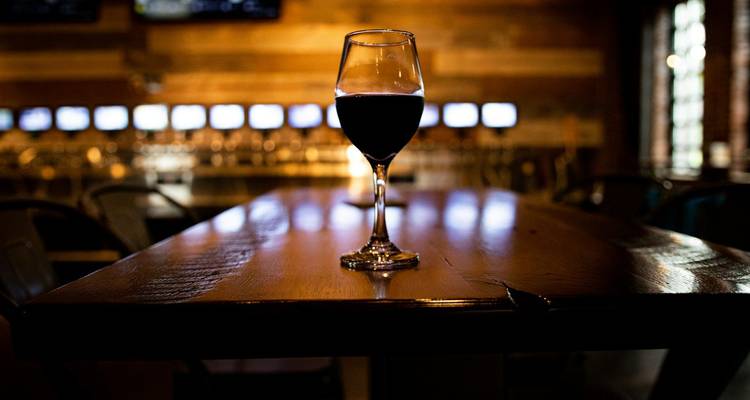 Una sola copa de vino tinto sobre una mesa de bar de madera pulida en una sala de cata con poca luz.