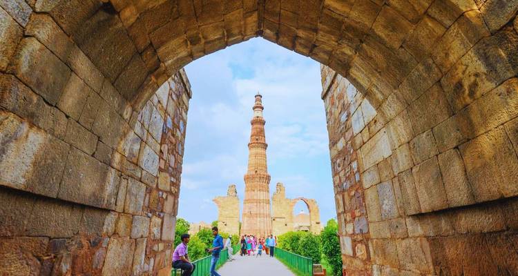 Visitantes enmarcados por un arco de piedra antigua contemplan la torre Qutub Minar que se alza en un día brillante