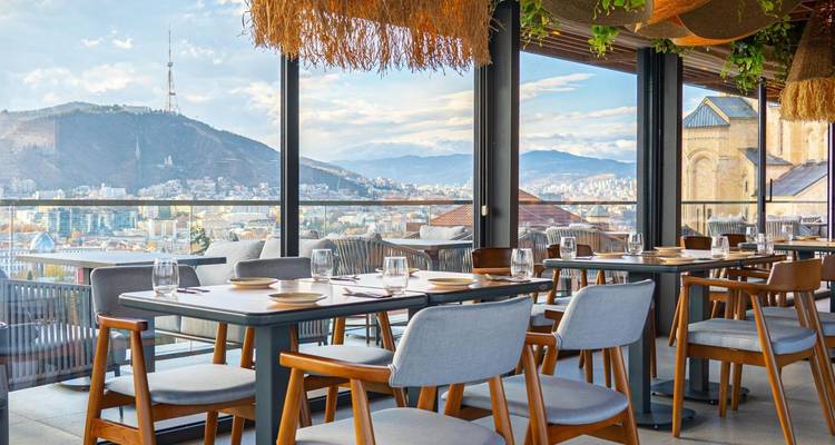 Helder restaurantinterieur met uitzicht op bergen en stadsskyline door glazen wanden, ingericht voor dineren.