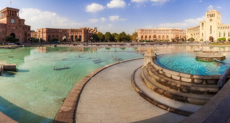 Amplio panorama de las piscinas de la fuente de la Plaza de la República y los edificios históricos de toba rosa bajo un cálido cielo vespertino en Ereván.