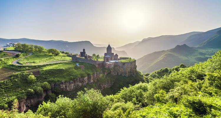 Historisches Kloster auf einer grünen Klippe, mit nebligen Bergen und Sonnenlicht bei Sonnenaufgang im Hintergrund.