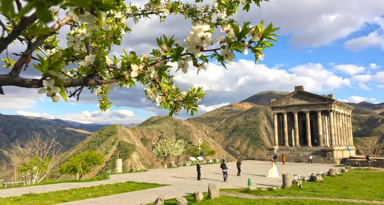 Pagan Garni Tempel, umrahmt von blühenden Baumzweigen mit Bergkulisse im Hintergrund.