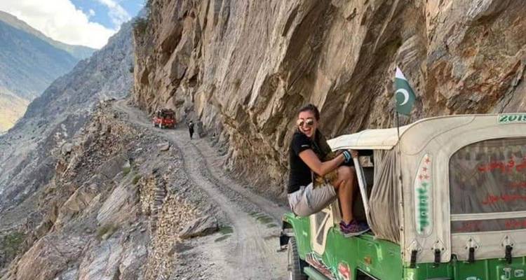 Adventurous traveller hanging out of green jeep on narrow cliffside road with Pakistani flag