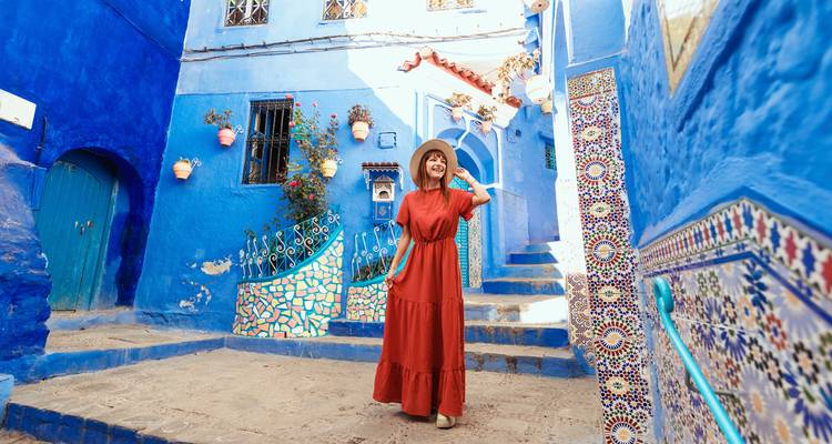 Smiling woman in flowing red dress stands in a picturesque blue street adorned with mosaic steps and potted plants in Chefchaouen.