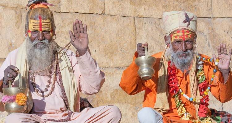 Twee kleurrijk beschilderde sadhu's zittend tegen een stenen muur, zegenend met geheven handen en rituele vaten vasthoudend.