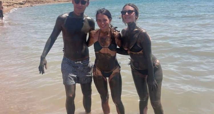 Trois amis couverts de boue de la mer Morte posant joyeusement dans de l'eau claire peu profonde.