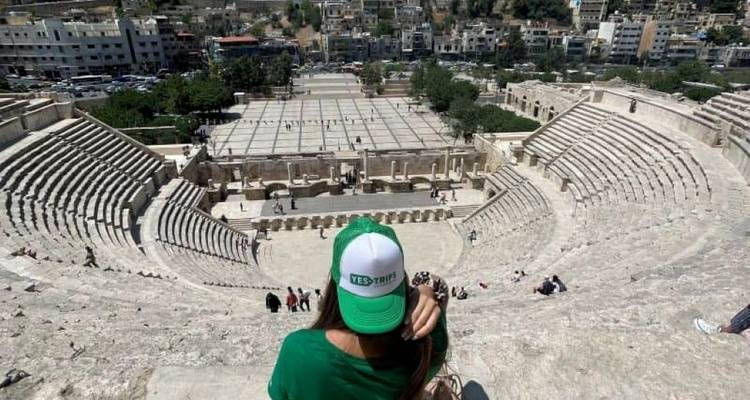Vue sur l'amphithéâtre romain antique d'Amman depuis les gradins élevés avec un voyageur portant une casquette verte au premier plan.