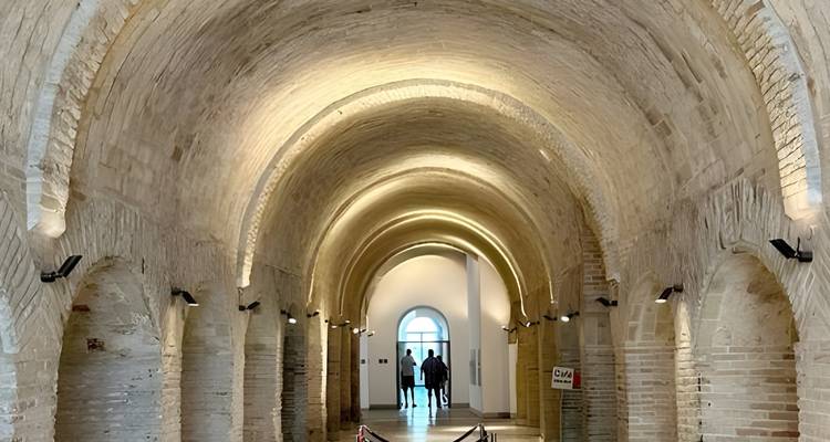 Stenen tongewelfde gang met zachte verlichting en twee bezoekers die naar het daglicht toe lopen.