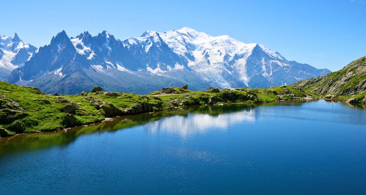 El lago alpino cristalino refleja el macizo del Mont Blanc cubierto de nieve bajo un cielo azul sin nubes.