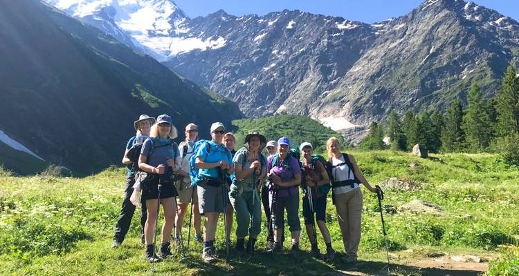 Grupo de senderismo posa en un prado alpino soleado con imponentes picos nevados al fondo.