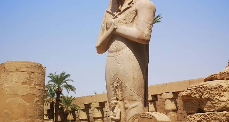 Eine hohe antike Statue eines Pharaos steht zwischen Palmen und verfallenen Säulen unter einem blauen Himmel.