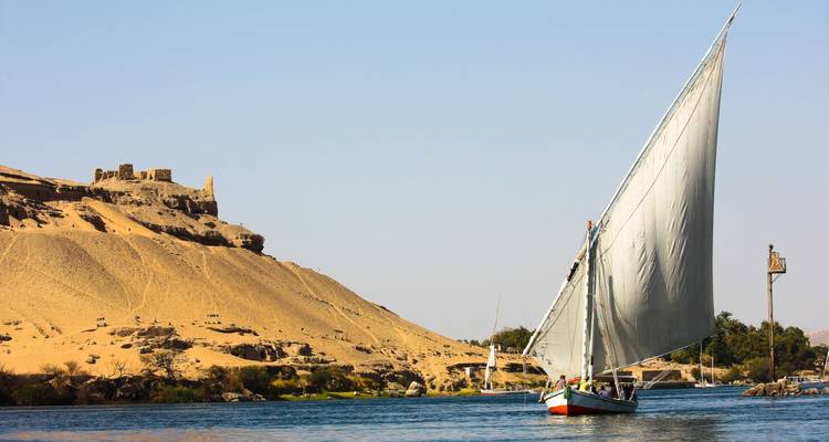 Eine traditionelle Feluke mit vollem weißen Segel gleitet den Nil entlang neben sandigen Hügeln und Ruinen.