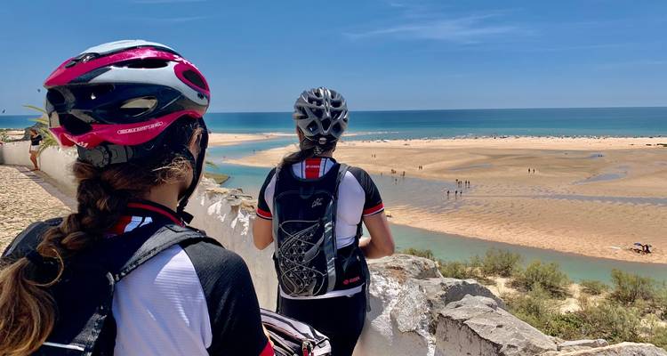 Zwei Radfahrer mit Helmen bewundern einen weitläufigen Sandstrand und türkisfarbenes Atlantikwasser.