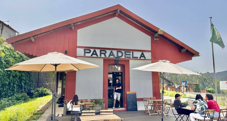 Radfahrer und Gäste entspannen sich vor einem kleinen roten Bahnhofscafé mit dem Schild "Paralela" inmitten von Grün.