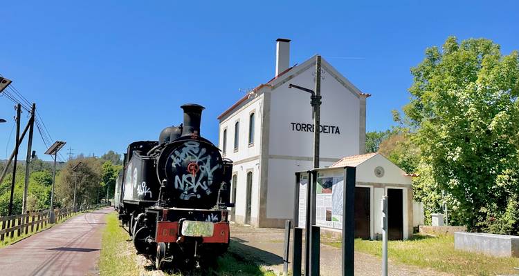 Eine mit Graffiti bedeckte Dampflokomotive ruht auf Schienen neben der weißen Station Torre deita unter blauem Himmel.