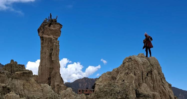 Una persona se encuentra en la cima de un pináculo rocoso erosionado en el paisaje lunar del Valle de la Luna contra un cielo azul profundo.