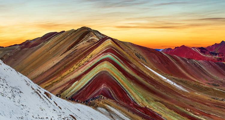 Montaña Arcoíris vibrante con bandas de rojo, amarillo y verde bajo un cielo dramático de amanecer, pequeños excursionistas en la cresta.