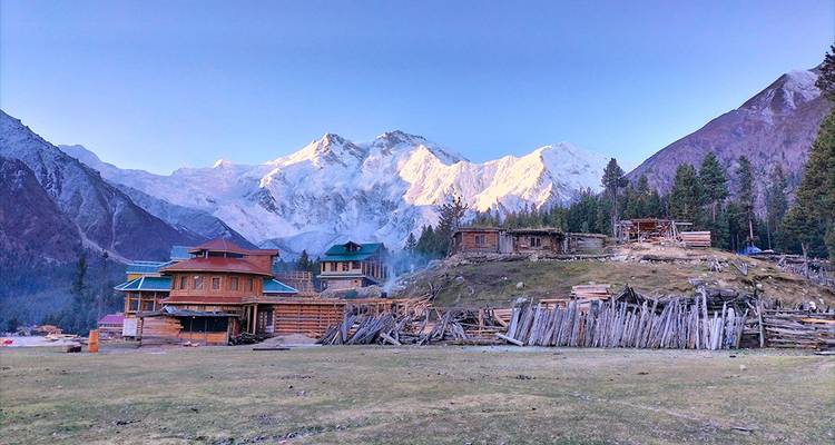 Chalets en bois et clôtures rustiques dans les Prairies des Fées avec le Nanga Parbat enneigé qui brille dans la lumière de l'aube.