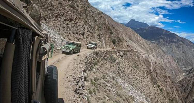 Convoi de jeeps naviguant sur un sentier de terre étroit au bord d'une falaise en haute montagne sous des nuages épars.
