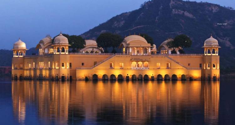 Het verlichte Jal Mahal paleis drijft op een kalm meer bij schemering met heuvels die zich als silhouet aftekenen erachter.